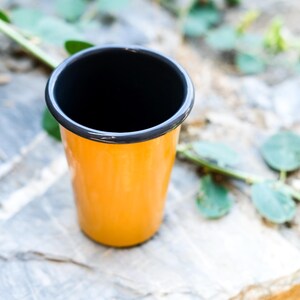 以下が含まれることがあります： 黒い縁取りのある黄色のエナメル製のマグカップ。マグカップは、緑の葉が背景にある薄い灰色の表面の上に置かれています。