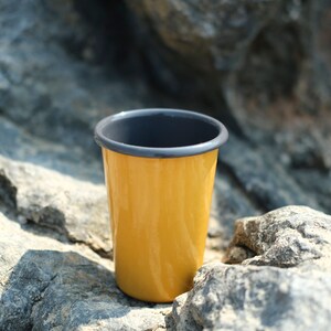 Close-up of a yellow enamel tumbler featuring an anthracite rim, placed on textured gray rocks. The sunlight reflects off the smooth surface of tumbler, emphasizing its glossy finish and the vibrant color contrast against the natural stone backdrop.