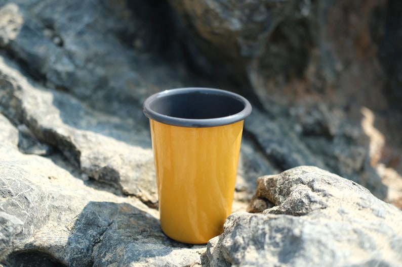 Close-up of a yellow enamel tumbler featuring an anthracite rim, placed on textured gray rocks. The sunlight reflects off the smooth surface of tumbler, emphasizing its glossy finish and the vibrant color contrast against the natural stone backdrop.