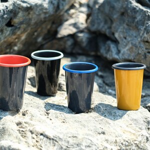 Handmade enamel tumblers in red, yellow, black, and blue, arranged on a rocky ground in an outdoor setting.