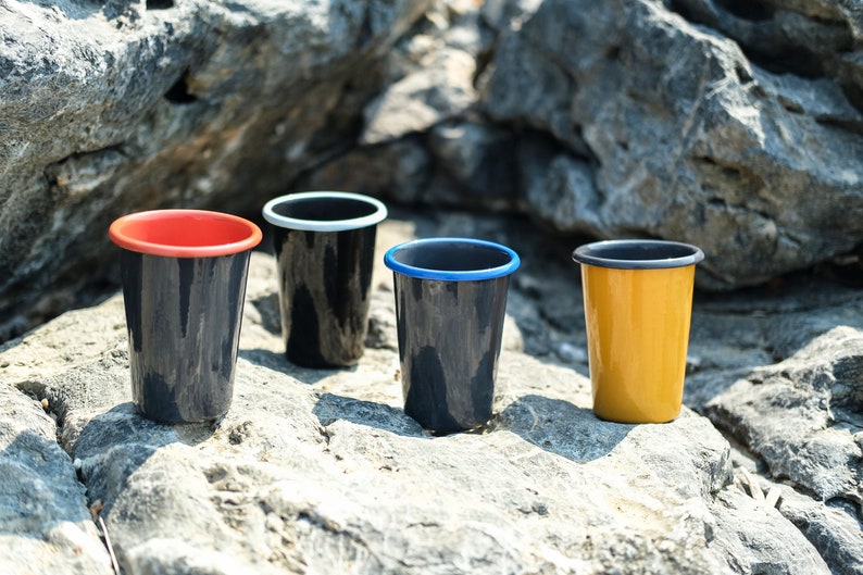 Handmade enamel tumblers in red, yellow, black, and blue, arranged on a rocky ground in an outdoor setting.