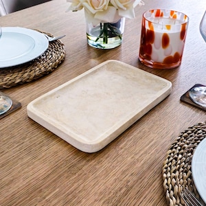 May include: A light beige rectangular serving tray made of marble with rounded edges. The tray is on a wooden table with a woven placemat and a white plate.