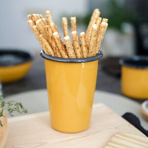 A rustic yellow enamel cup with an anthracite rim holds golden sesame breadsticks. The table is set with an emanel mug, an enamel bowl, and a glass bowl of chocolate adds a savory touch, creating a warm, inviting snack scene.