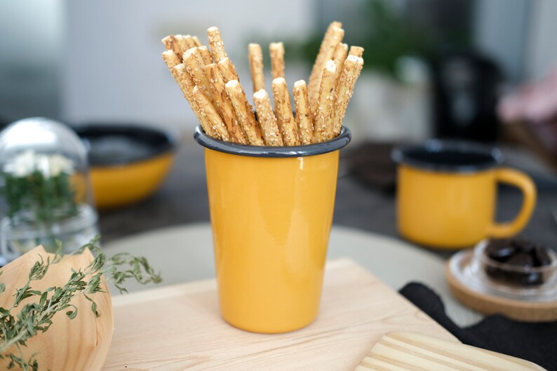 A rustic yellow enamel cup with an anthracite rim holds golden sesame breadsticks. The table is set with an emanel mug, an enamel bowl, and a glass bowl of chocolate adds a savory touch, creating a warm, inviting snack scene.