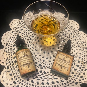 Peut inclure: Deux petites bouteilles en verre avec des compte-gouttes, &eacute;tiquet&eacute;es "Ravenwood Apothecary Ashwagandha" et contenant un liquide fonc&eacute;. Les bouteilles sont sur une dentelle blanche en crochet avec un verre de liquide ambr&eacute; en arri&egrave;re-plan.