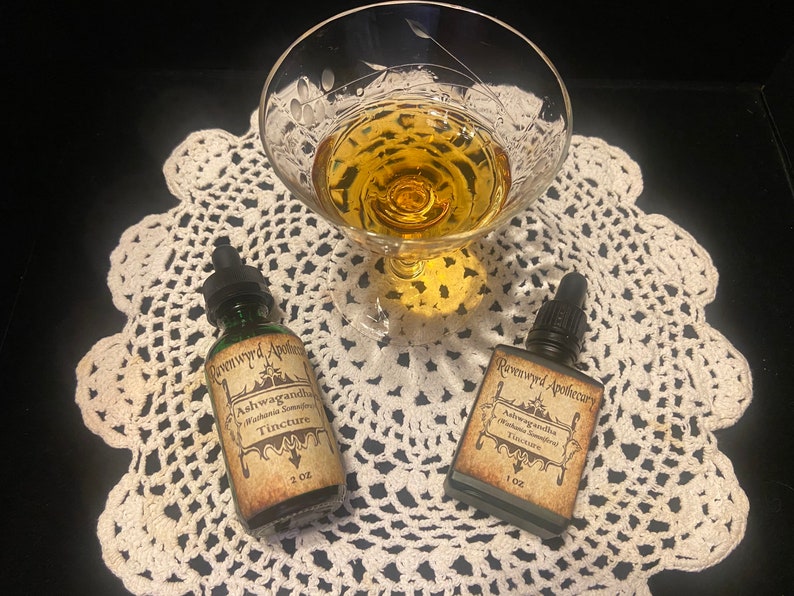 Peut inclure: Deux petites bouteilles en verre avec des compte-gouttes, &eacute;tiquet&eacute;es "Ravenwood Apothecary Ashwagandha" et contenant un liquide fonc&eacute;. Les bouteilles sont sur une dentelle blanche en crochet avec un verre de liquide ambr&eacute; en arri&egrave;re-plan.