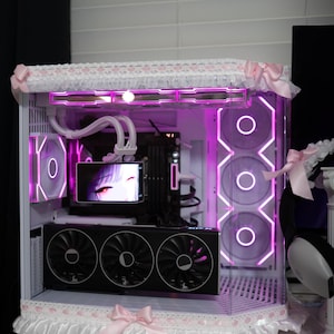 May include: A custom PC case with a white and pink design. The case has a clear side panel, revealing internal components lit by pink LED lights. The case is decorated with frills and bows, giving it a unique and decorative look. The case is on a white surface.