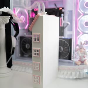 May include: A white, house-shaped computer case with a pink roof and window frames. The case has three rows of windows and a chimney. The background features a computer with pink lighting and a decorative trim.