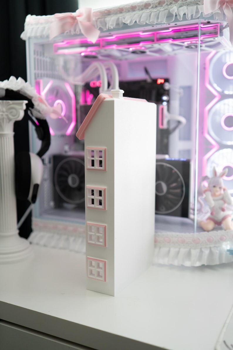 May include: A white, house-shaped computer case with a pink roof and window frames. The case has three rows of windows and a chimney. The background features a computer with pink lighting and a decorative trim.