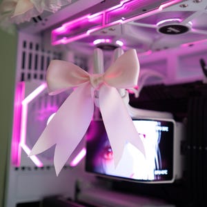 May include: A white computer case with pink LED lighting and a large pink bow. The case has a decorative trim with lace and additional pink bows. Computer components are visible inside the case.