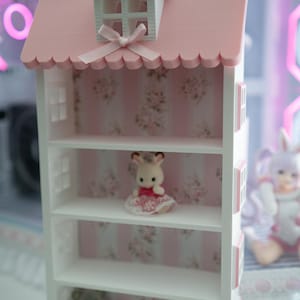 May include: A white dollhouse-style shelf unit with a pink roof and floral wallpaper backing. The roof has a small dormer window and a decorative bow. The shelves display small figurines, including a bunny in a dress and a teddy bear.