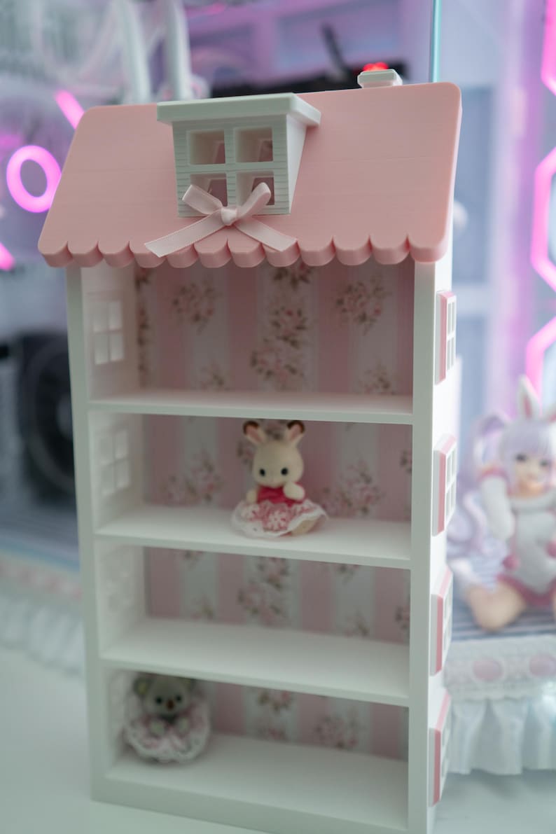 May include: A white dollhouse-style shelf unit with a pink roof and floral wallpaper backing. The roof has a small dormer window and a decorative bow. The shelves display small figurines, including a bunny in a dress and a teddy bear.