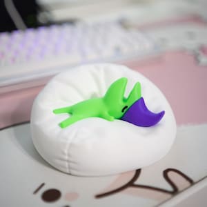 May include: A quirky green and purple character figurine lounging on a small white beanbag. The figurine has a distinctive design and a relaxed posture. The beanbag sits on a pink and white surface, suggesting a playful or decorative theme.