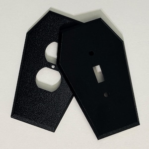 May include: Two black, coffin-shaped light switch covers. One cover has openings for a standard outlet, the other has a single switch opening. The covers are made of a textured material.