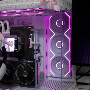 以下が含まれることがあります： 白とピンクを基調としたカスタムメイドのコンピューターケース。ケースはガラスパネル、ピンクのLED照明、装飾トリムを備えています。デザインには、円形のライトが付いた3つの大きなファンとピンクのリボンが含まれています。小さな猫の形をしたアイテムが前景にあります。