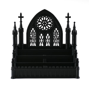 Puede incluir: Un expositor negro de varios niveles con un diseño inspirado en una catedral, que presenta un rosetón central, arcos apuntados y cuatro agujas laterales rematadas con cruces. La estructura incluye tres estantes horizontales para organizar.