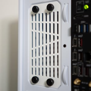 May include: Close-up of a white computer case with a rectangular vent cover secured by four black screws. The vent cover has vertical slots for airflow. A portion of the motherboard is visible.