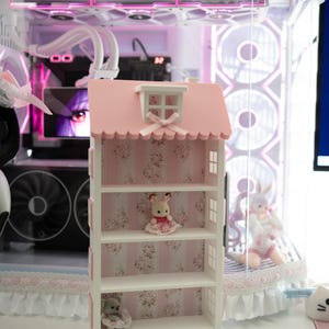 May include: A pink and white dollhouse-shaped shelf unit with floral wallpaper backing. The shelf is decorated with small figurines, including a bunny and a bear. The background features a computer with pink lighting and decorative elements.
