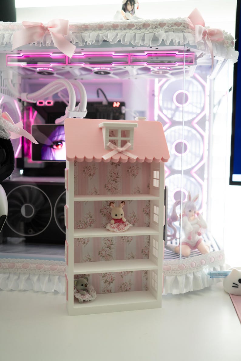 May include: A pink and white dollhouse-shaped shelf unit with floral wallpaper backing. The shelf is decorated with small figurines, including a bunny and a bear. The background features a computer with pink lighting and decorative elements.
