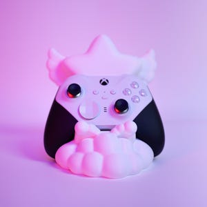 May include: A white and black wireless game controller rests in a white stand shaped like a stylized cloud with wings. The controller features a central Xbox logo, two black analog sticks, a circular directional pad, and various buttons on the face.