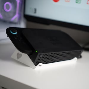 May include: A black gaming console with a controller attached, resting on a white stand with a heart design. The console has a perforated side and a green power indicator. The background includes a computer monitor and a purple-lit fan.