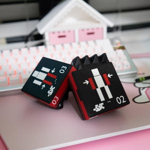 May include: Two black and red square-shaped game pieces with white and red graphics and the numbers 02 and 03. The pieces are on a pink surface with a white keyboard and a small white house in the background.