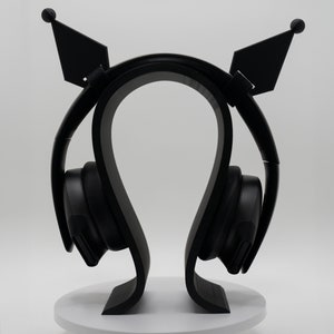 May include: Black headphones with a matching black stand. The stand has a unique design with curved supports and decorative ear-shaped accents. The headphones are resting on a white circular base, creating a sleek and modern aesthetic.