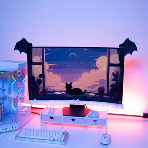 May include: A computer monitor featuring a digital illustration of a cat looking out a window at a night sky, adorned with black bat wings on the top corners and a white ruffled trim along the bottom edge, set on a white desk with a keyboard and mouse.