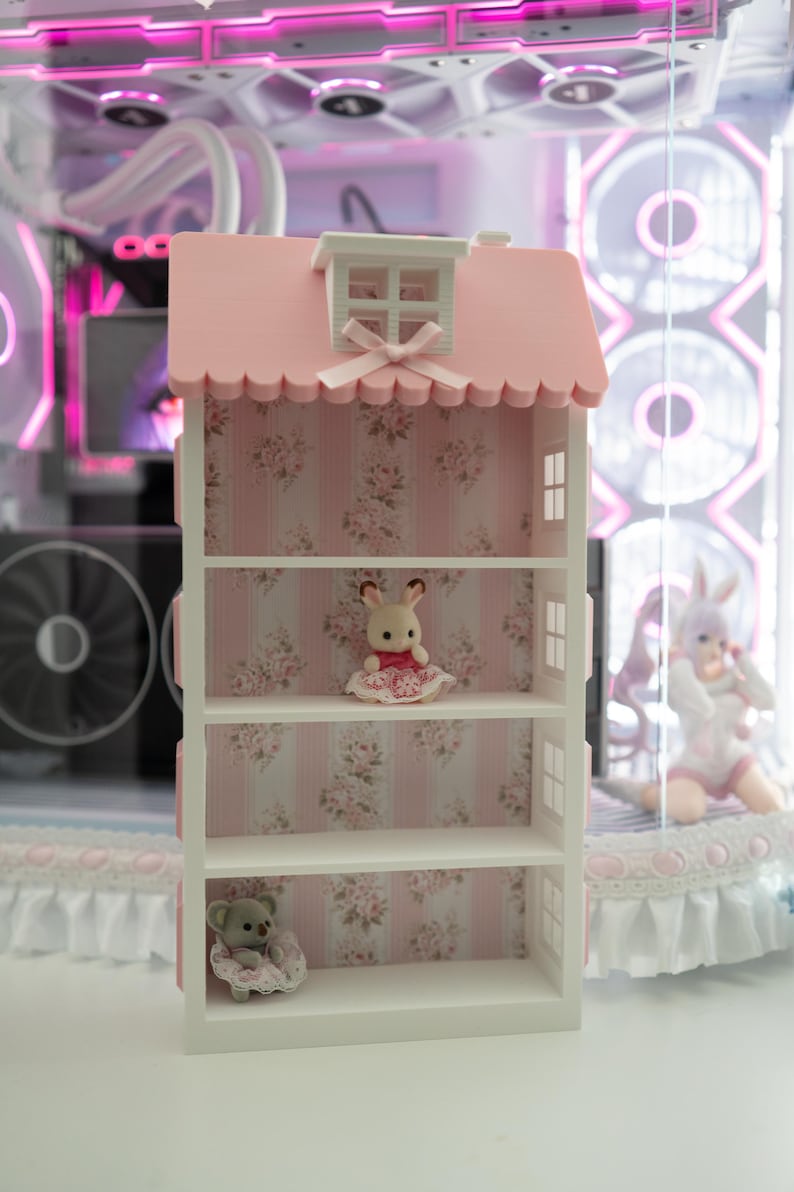 May include: A white and pink dollhouse-style shelf unit with three shelves. The roof is pink with a scalloped edge and a small window. The interior features floral wallpaper and includes a small bunny and a teddy bear figurine.
