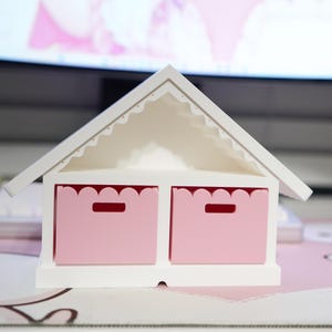 May include: A white wooden house-shaped storage unit with a scalloped roof and two pink drawers. Each drawer has a rectangular handle cutout. The unit sits on a light pink surface.