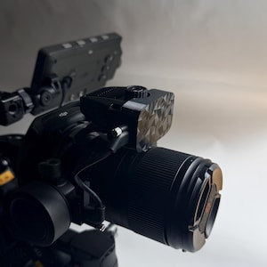 May include: A professional black camera with a large lens and attached accessories. The camera has a geometric patterned top piece and is set against a neutral background. The lens has a silver ring.