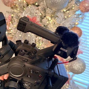 May include: A black video camera with a Rode microphone attached. The camera has various buttons and dials, including a record button. The background features a decorated Christmas tree with pink and silver ornaments.