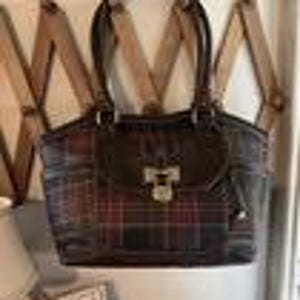 May include: A dark brown and red plaid handbag with a top handle and a rectangular shape. The bag has a flap closure with a silver-colored clasp. The bag is hanging on a wooden wall-mounted rack.