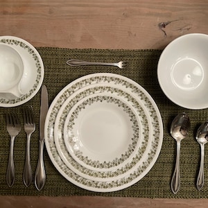 May include: A set of white dinnerware with a green floral pattern. The set includes a dinner plate, a salad plate, a bowl, a cup, and a saucer. There are also four silver forks, two silver spoons, and a silver knife on a green placemat.