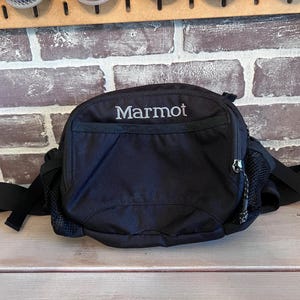 May include: Black Marmot fanny pack with a zippered main compartment and a front pocket. The pack has a mesh side pocket and adjustable straps. The Marmot logo is embroidered in white.