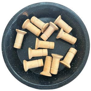 May include: A collection of unfinished wooden peg dolls with a flared top and cylindrical body, arranged in a speckled black bowl. The peg dolls are made of light-colored wood.