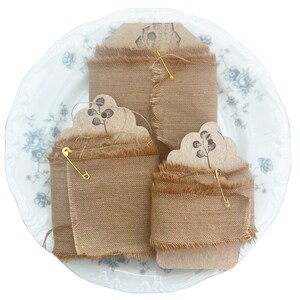 May include: Three rolls of tan burlap ribbon with frayed edges are displayed on a white plate with a floral design. Each roll is secured with a gold safety pin and a decorative tag with a botanical print.