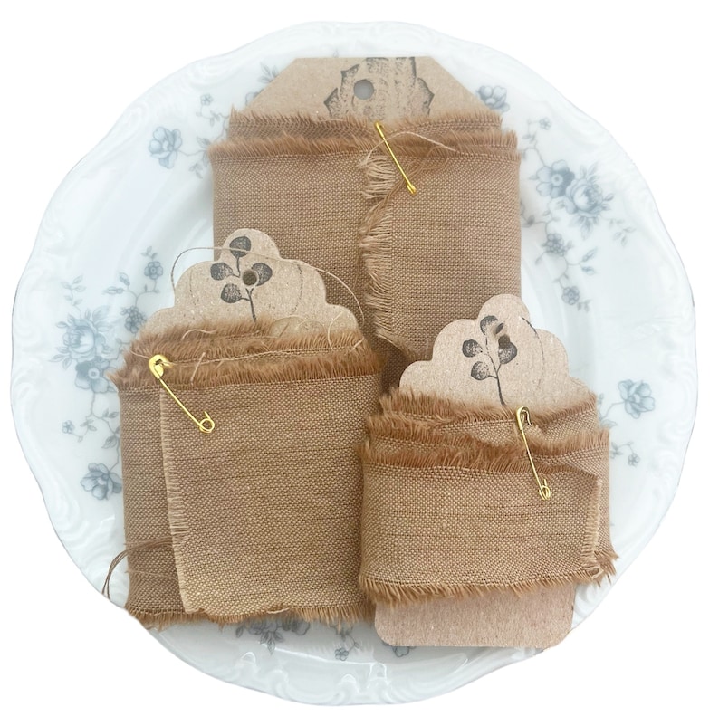 May include: Three rolls of tan burlap ribbon with frayed edges are displayed on a white plate with a floral design. Each roll is secured with a gold safety pin and a decorative tag with a botanical print.