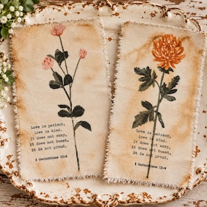 May include: Two rectangular fabric pieces with frayed edges, featuring tea-stained backgrounds. One displays pink flowers, the other an orange flower. Both include the black printed text: Love is patient, love is kind. It does not envy, it does not boast, it is not proud. 1 Corinthians 13:4.