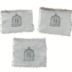 May include: Three square burlap coasters with frayed edges. Each coaster features a gray illustration of a birdcage with two birds inside. The coasters are a neutral color, suitable for various home decor styles.