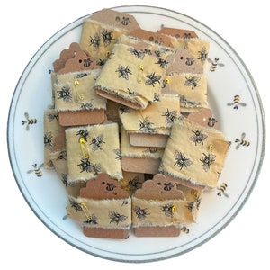 May include: A collection of beige fabric ribbon spools with a black bee print. Each spool is wrapped around a brown cardboard tag with a gold safety pin.