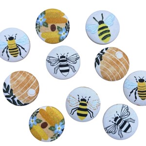 May include: A collection of ten round fabric buttons with bee-themed designs. The buttons feature images of bees, honeycombs, and floral patterns in yellow, black, white, and blue. The buttons are arranged on a white background.