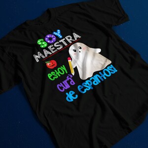 Op de afbeelding: Zwart T-shirt met een kleurrijk grafisch ontwerp met een spook, een appel, een potlood en de tekst "Soy Maestra, Estoy Cura de Espanhol".