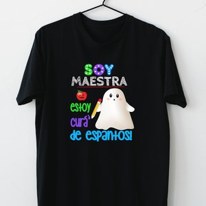 Op de afbeelding: Zwart T-shirt met een kleurrijk grafisch ontwerp met een spook dat een potlood vasthoudt en de tekst "Soy Maestra, estoy cura de espantos!"
