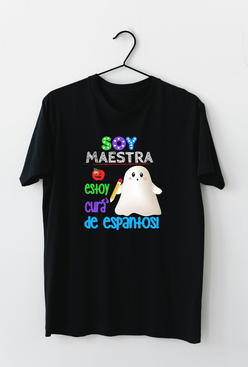 Op de afbeelding: Zwart T-shirt met een kleurrijk grafisch ontwerp met een spook dat een potlood vasthoudt en de tekst "Soy Maestra, estoy cura de espantos!"