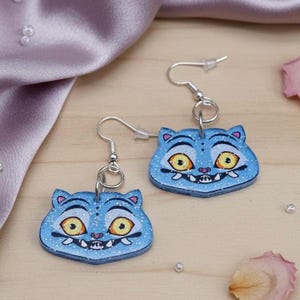 Pode incluir: Um par de brincos azuis em forma de gato com ganchos prateados. Cada brinco apresenta um rosto de gato de desenho animado com olhos amarelos, um sorriso com dentes brancos e detalhes rosa. Os brincos têm um acabamento brilhante.