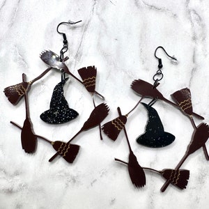 May include: A pair of witch hat earrings with a black hat and brown broomstick accents. The earrings are made of acrylic and have a glitter finish.