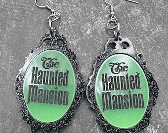 Könnte beinhalten: Ein Paar silberne Ohrringe mit einem grünen, glitzernden ovalen Anhänger, auf dem "The Haunted Mansion" steht.