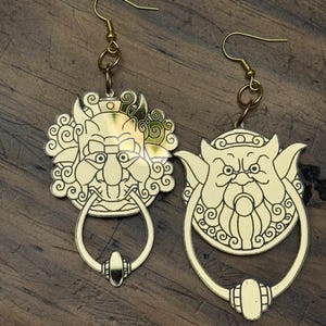 May include: A pair of gold-tone earrings featuring a detailed design of a mythical creature's face with a knocker-style ring. The earrings are a unique and eye-catching accessory.