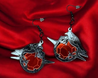 May include: A pair of silver earrings featuring a stylized design of a bird holding a red gemstone. The earrings are on a red satin background.